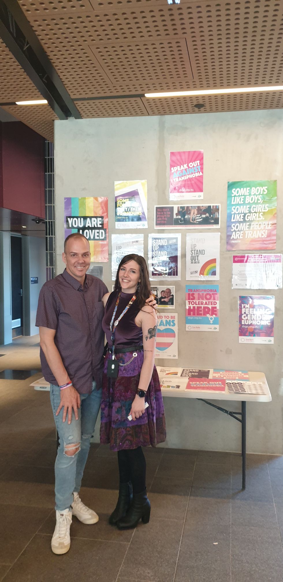 Two people smiling at the camera with flyers and posters in the background