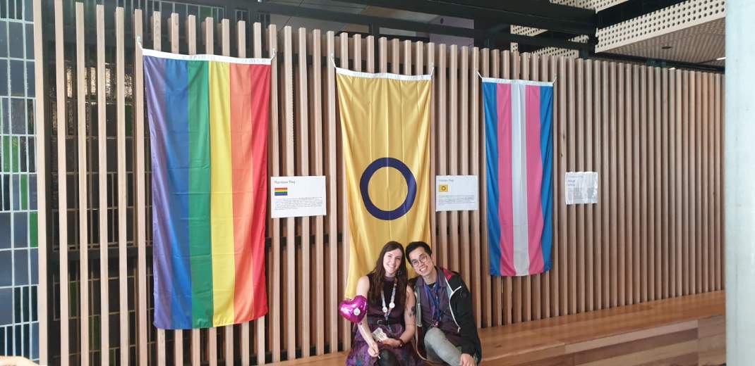 Two people sitting in front of a rainbow flag, intersex flag and transgender flag.